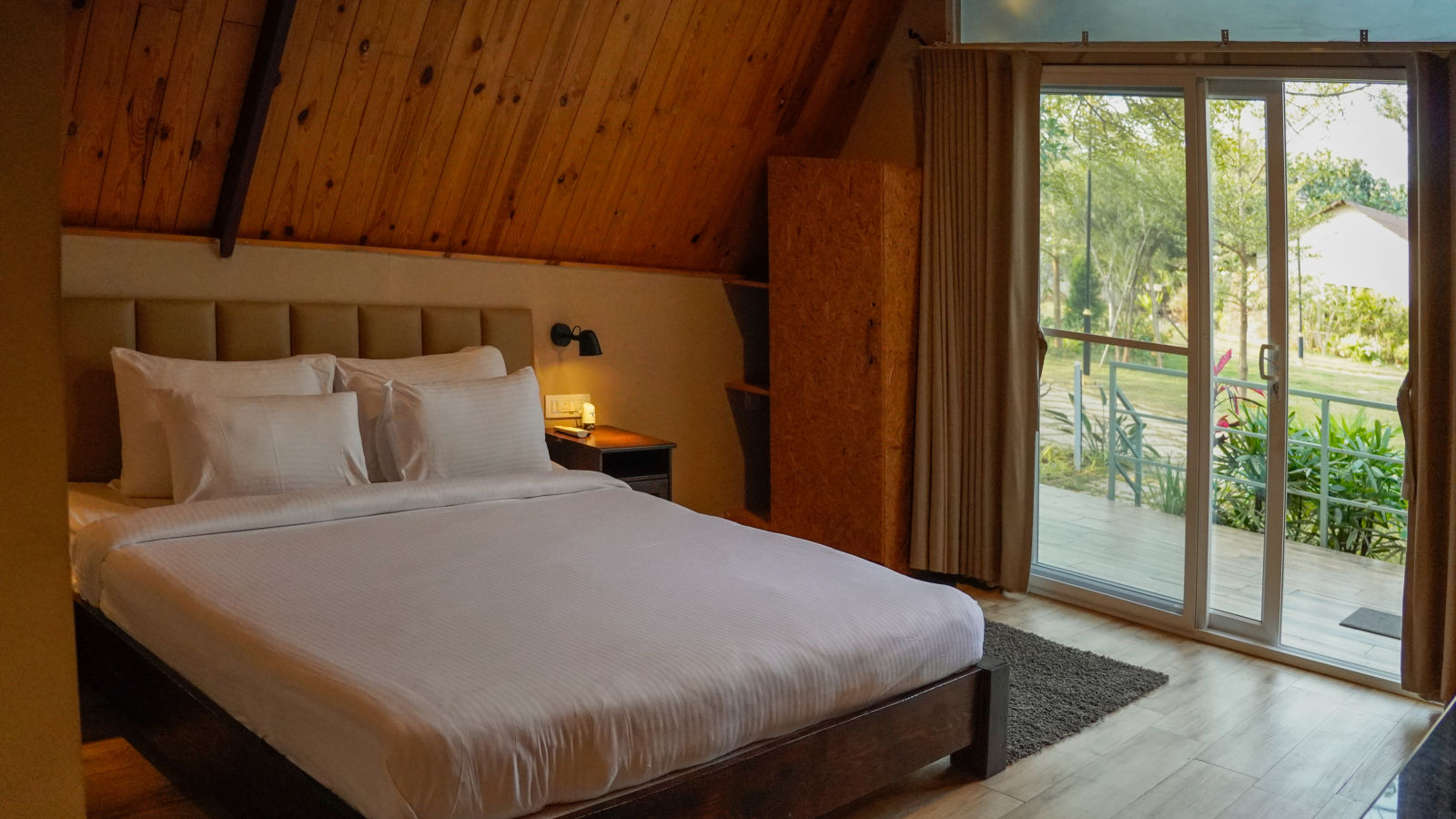 An image of the Aradhya room with a king size bed with a bed side table and an expansive slider door overlooking at the lawn - Trance Veechika Resort