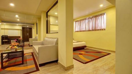 A hotel suite's living space featuring cream sofas, a colorful rug, and a bedroom view through a large glass window. 