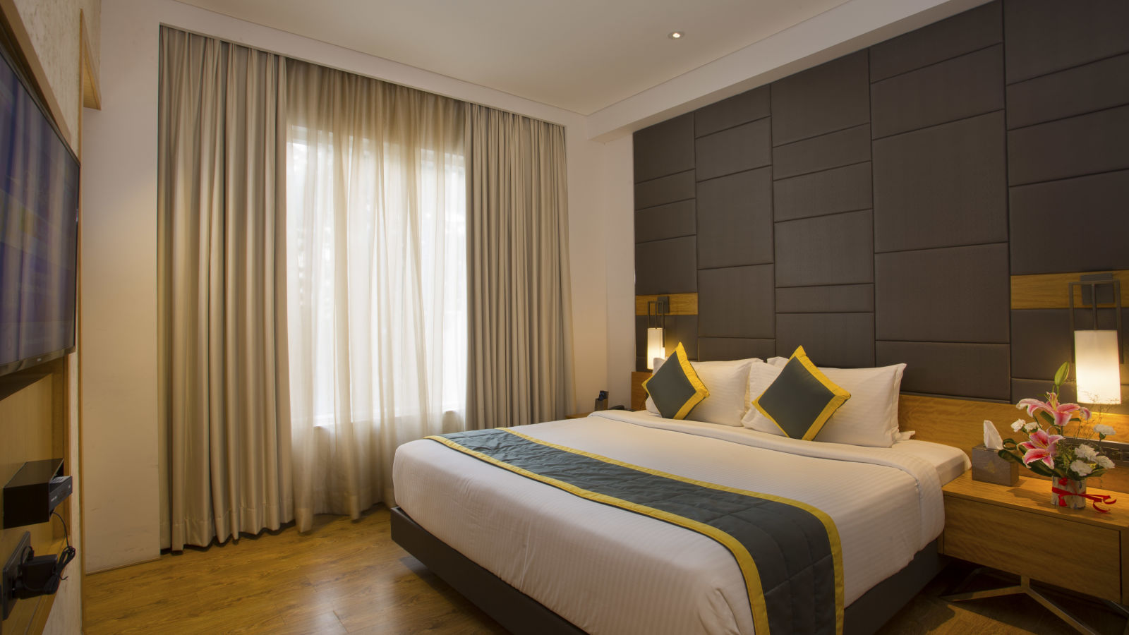 a bedroom with a large bed opposite a television mounted on the wall and side drawers on each side - Hotel Fountain Tree By TGI - JP Nagar, Bangalore