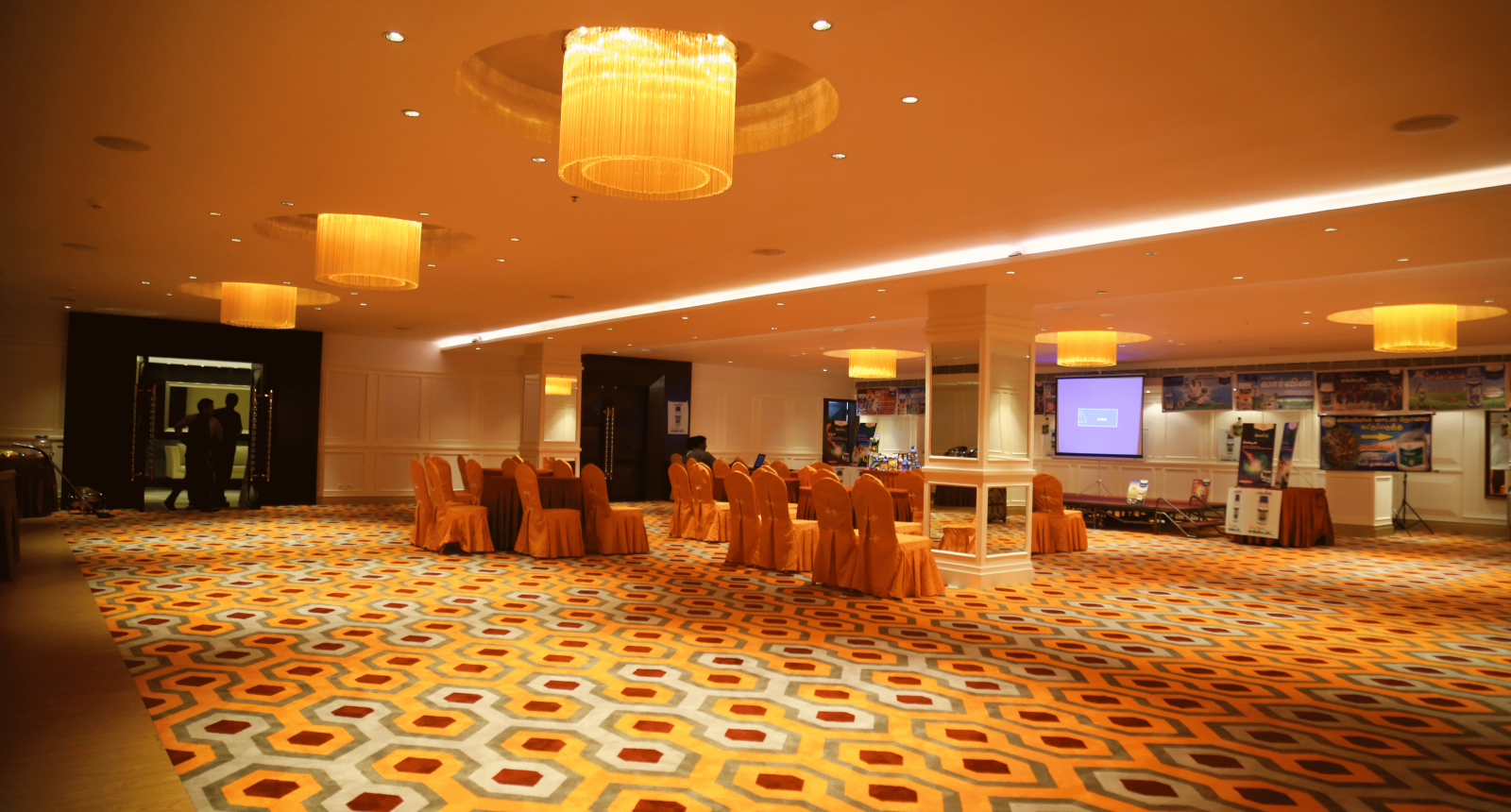 A banquet hall with orange chair covers and a central projection screen, under warm yellow lighting - TGI Grand Fortuna, Hosur