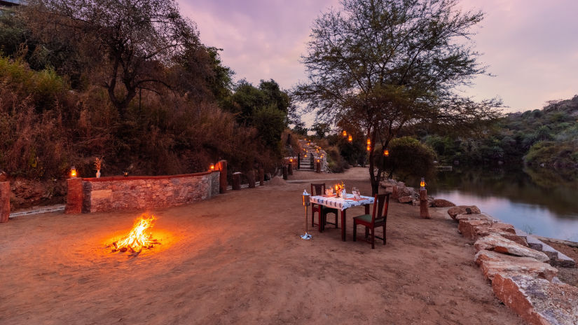 a table for two set up next a lake and coconut trees with a campfire as well - Chunda Shikar Oudi, Udaipur