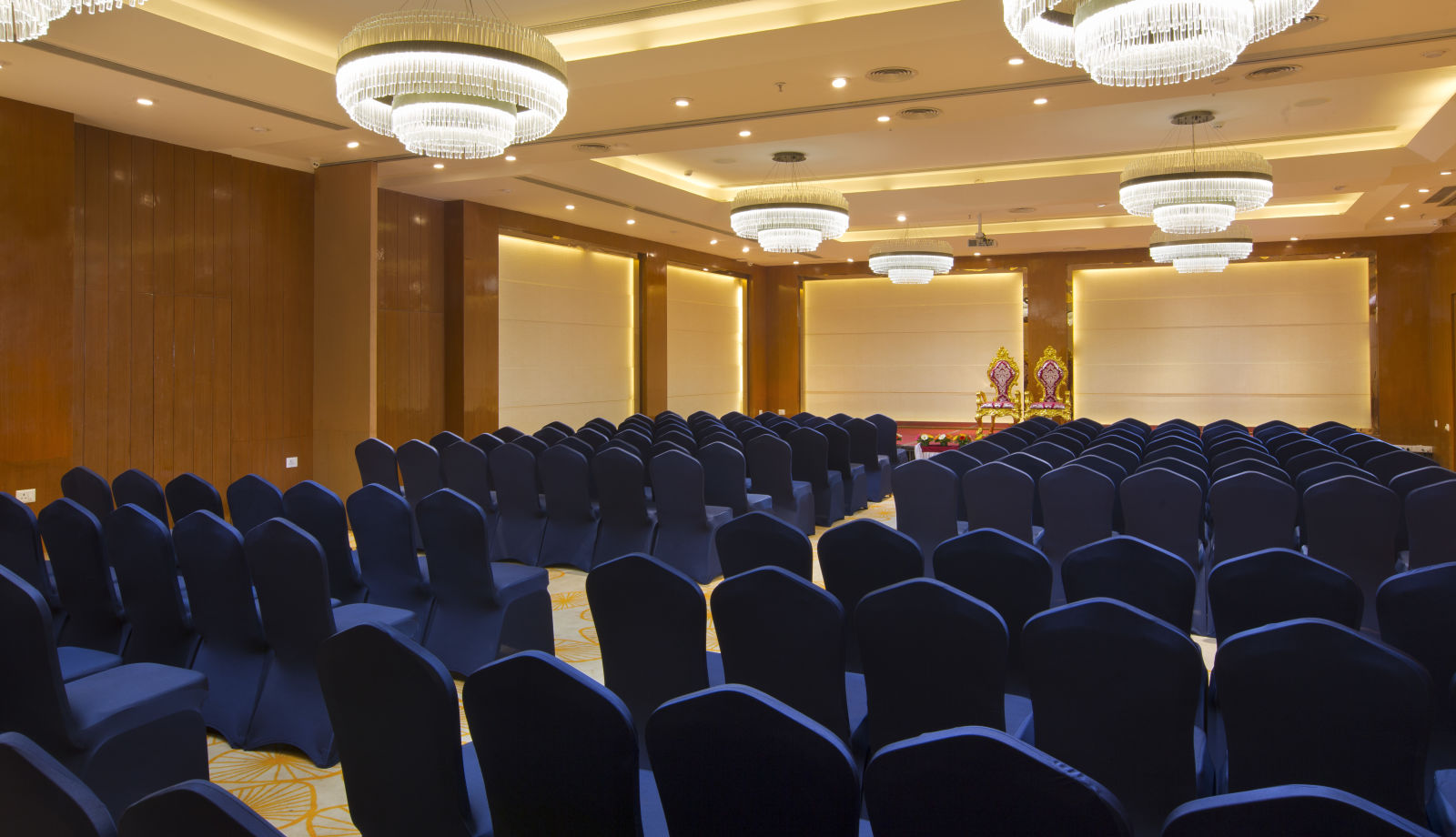 Another view of a spacious conference hall with rows of blue chairs facing a stage with golden figurines, illuminated by large chandeliers - Hotel Southend By TGI - Bommasandra, Bangalore