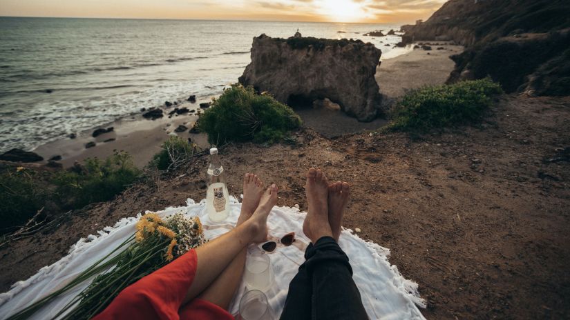 a lower angle shot of a couple resting on the beach after a picnic with a sunset as a backdrop