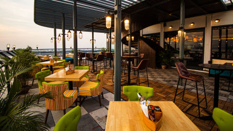 A scenic rooftop restaurant with colorful chairs, wooden tables, and a view of the sunset over a river.