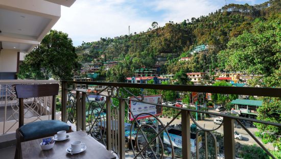 alt-text A building's balcony adorned with a chair, a coffee table bearing two tea cups and a bowl, all with a view overlooking a picturesque mountain town - Vinsober Monal, Nainital