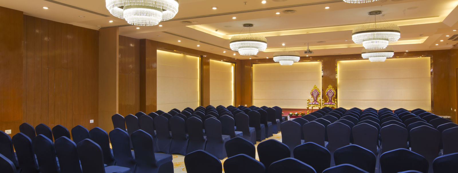 Another view of a spacious conference hall with rows of blue chairs facing a stage with golden figurines, illuminated by large chandeliers - Hotel Southend by TGI - Bommasandra, Bangalore