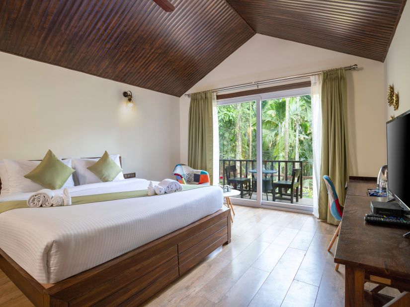 A hotel bedroom with a large bed with a green runner, a television and a balcony with the view of the garden - Stone Wood Nature Resort, Gokarna