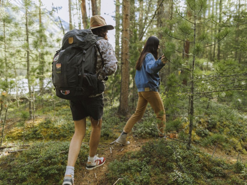 A couple walking on a trekking trail next to trees carrying a large backpack