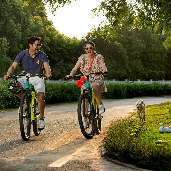 a couple cycling on a stone pathway next to nature and trees with the sun shining from behind them at this Romantic Getaways Delhi - Heritage Village Resort & Spa, Manesar