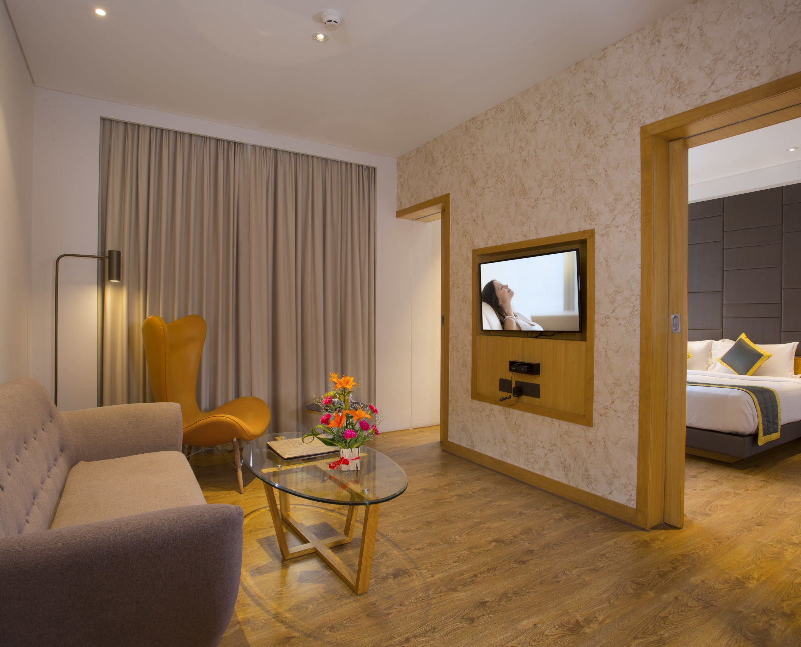 view of a living room with sofas and a small table, all facing a television mounted on the wall and a door leading to the bedrrom on the right - Hotel Fountain Tree By TGI - JP Nagar, Bangalore