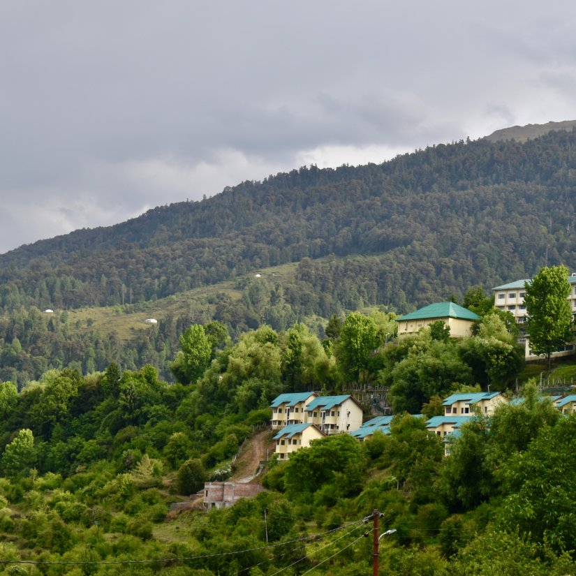 a view from afar of building amidst nature above a small hill with mountains in the background