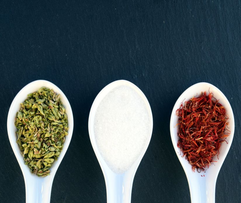 Three spoons on a dark surface, one with green herbs, one with white salt, and one with red spice.