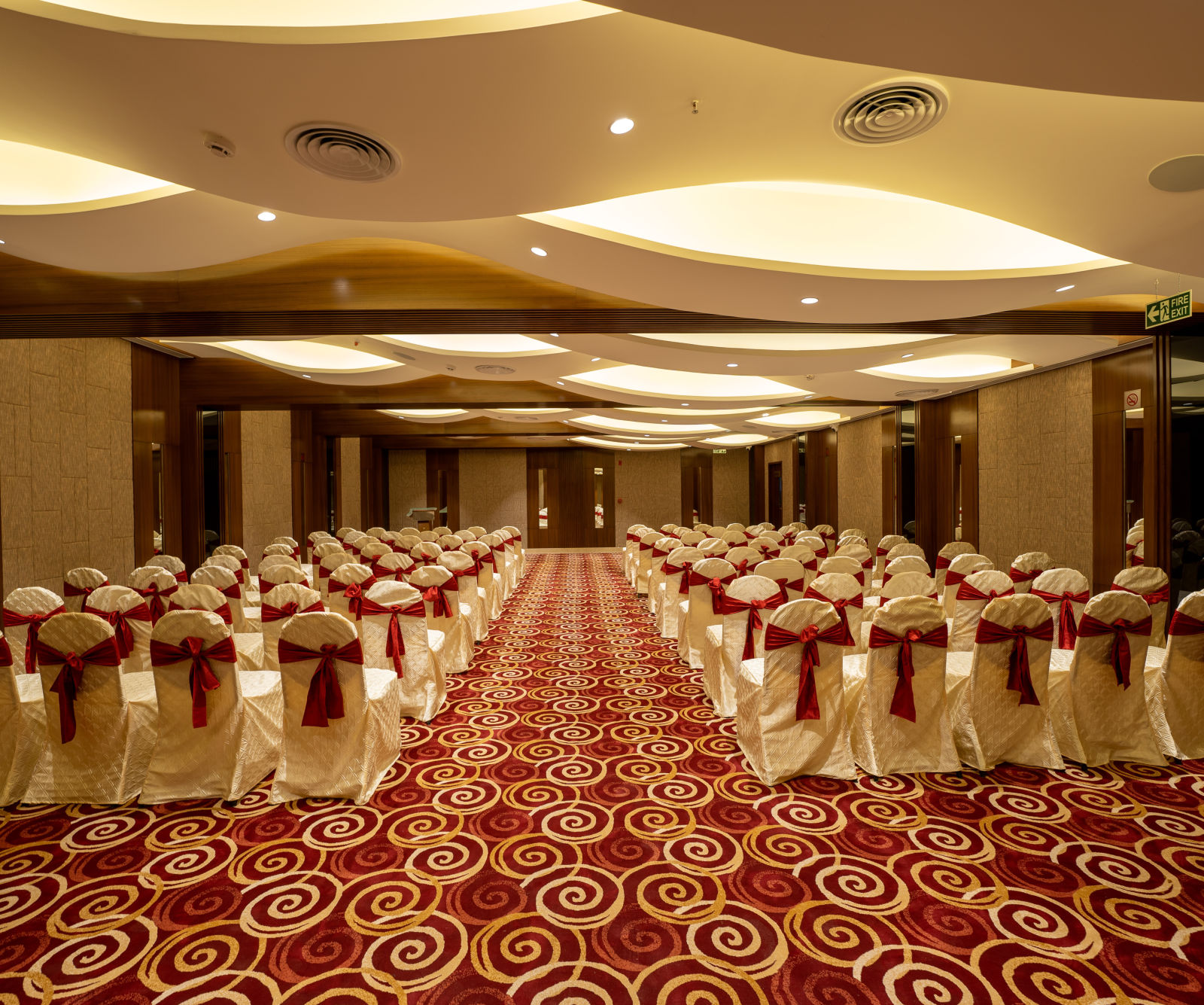 Large banquet hall set for an event with chairs covered in white and a red carpet.-The President Hotel, Kumara Park