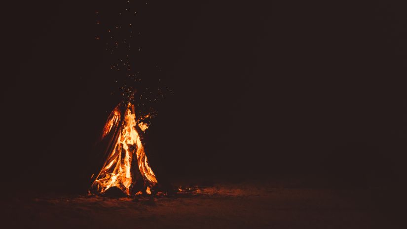 a raging bonfire made from long logs on a beach with darkness in the background