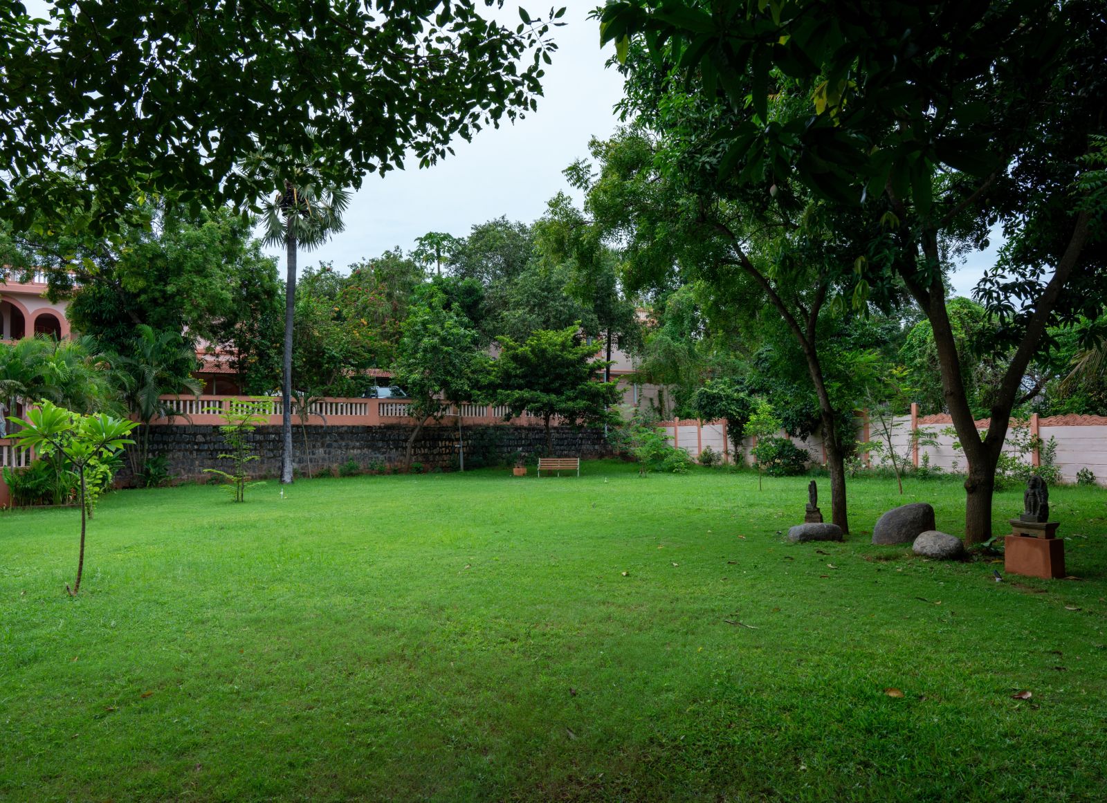 A lush green garden with scattered trees, statues, and a bench, surrounded by a low wall and shaded by large trees.