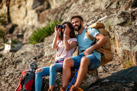 alt-text A couple sitting on rocks with hiking gear. One is using binoculars while the other looks on, set against a rocky and green background suggesting an exploration theme.