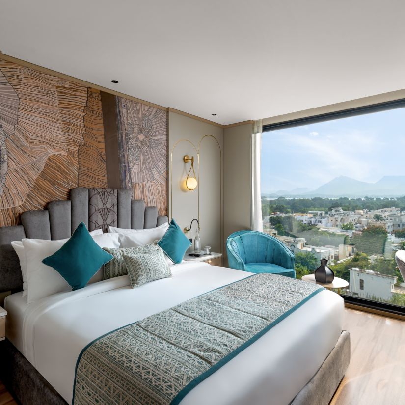A king bed inside a room at Parallel hotel with a view of the city from the floor to ceiling glass window , with all the modern aminities , lamp at the side table  and a beautiful scenic beauty at Parallel Hotel  Udaipur