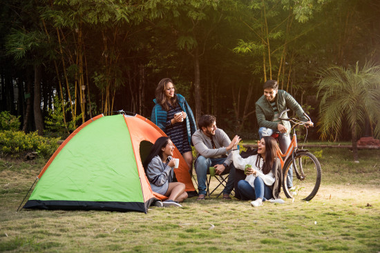 alt-text  A group of six people, including adults and teenagers, are gathered around a campsite with a green and orange tent. They are enjoying a meal and drinks together, with bicycles parked nearby.