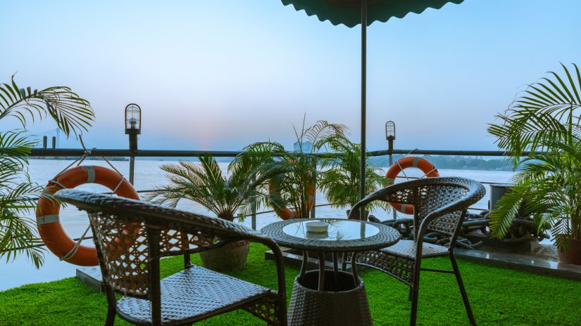 Relaxing outdoor balcony setting with rattan chairs and a table, overlooking a serene waterfront at dusk, surrounded by lush potted plants. - Polo Floatel Kolkata