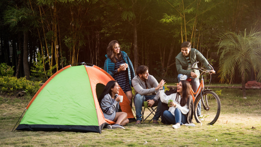  A group of six people, including adults and teenagers, are gathered around a campsite with a green and orange tent. They are enjoying a meal and drinks together, with bicycles parked nearby.