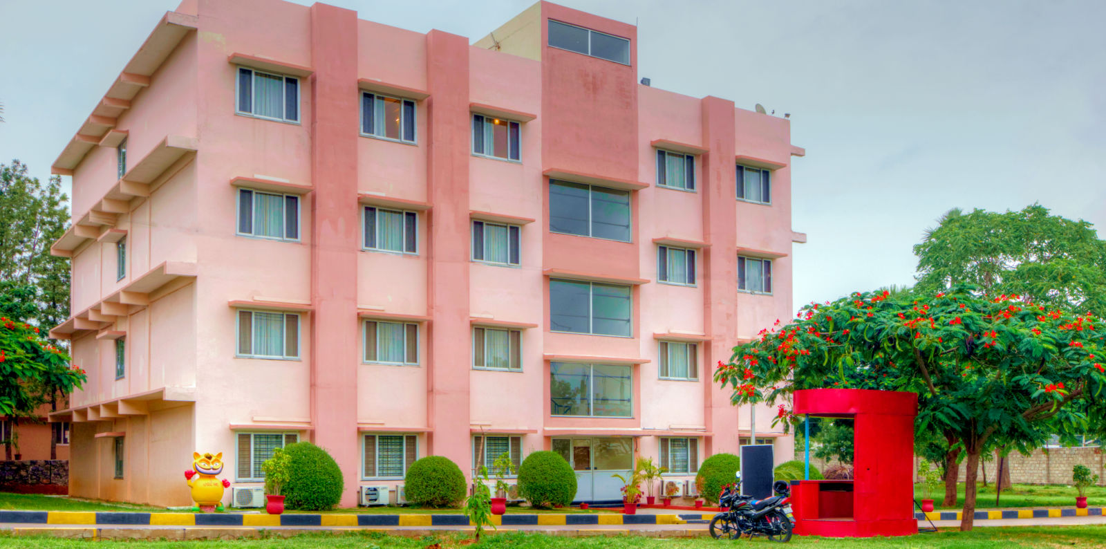 A vibrant exterior shot of a pink hotel building with greenery and a red security booth in the foreground. Greens Inn - Cozy Accommodations