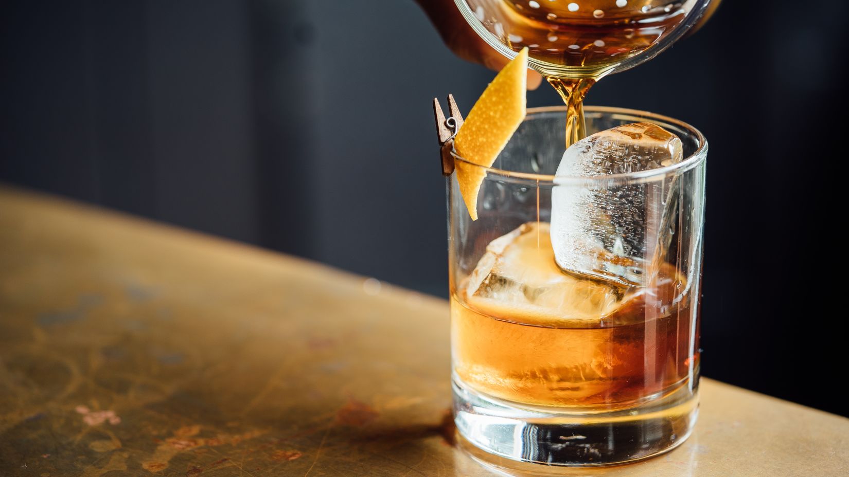 a person pouring a drink on a glass with an ice cube that is placed on a desk