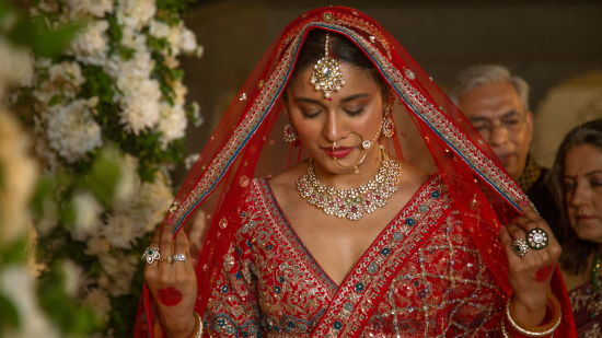 alt-text A bride in traditional indian attire, featuring a red lehenga and veil, adorned with intricate gold jewelry, looks down modestly during a wedding ceremony.