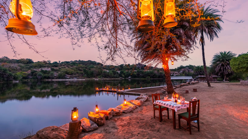 a table for two set up next to the lake and lamps hung on the tree next to the table - Chunda Shikar Oudi, Udaipur