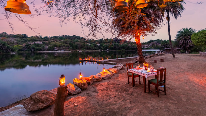 a table for two set up next to the lake and lamps hung on the tree next to the table - Chunda Shikar Oudi, Udaipur