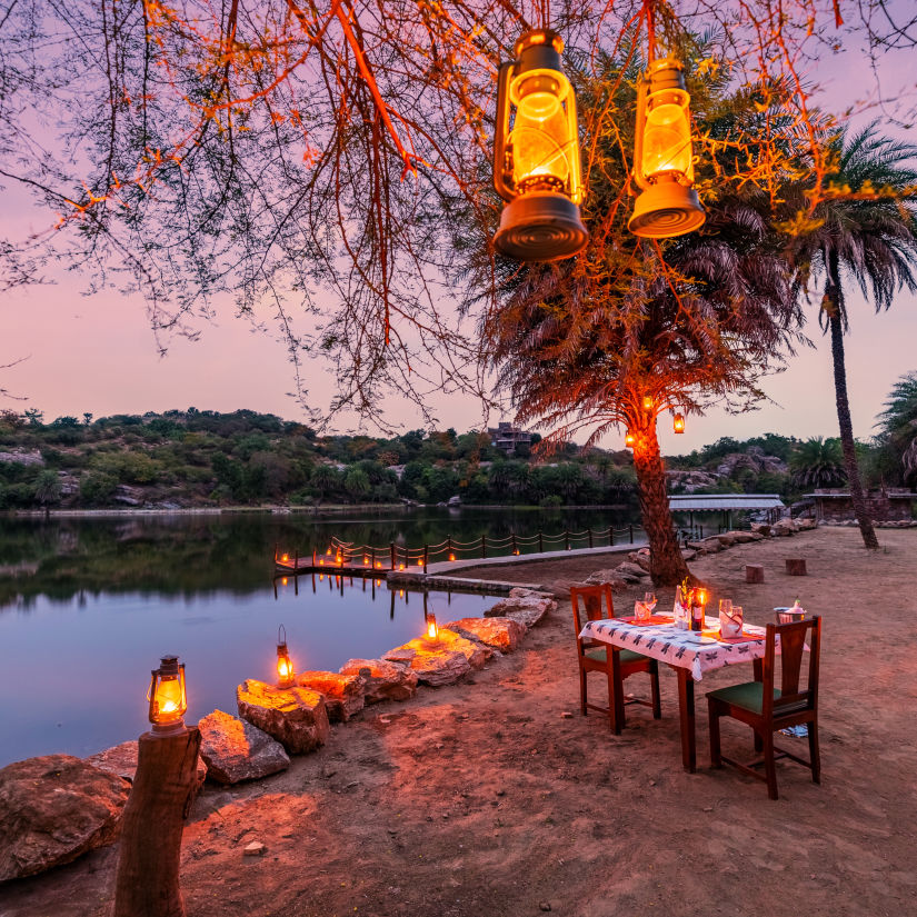 a table for two set up next to the lake and lamps hung on the tree next to the table - Chunda Shikar Oudi, Udaipur