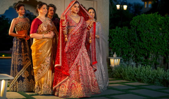 alt-text Four women in traditional indian sarees pose together at dusk, with soft lighting and a garden in the background.