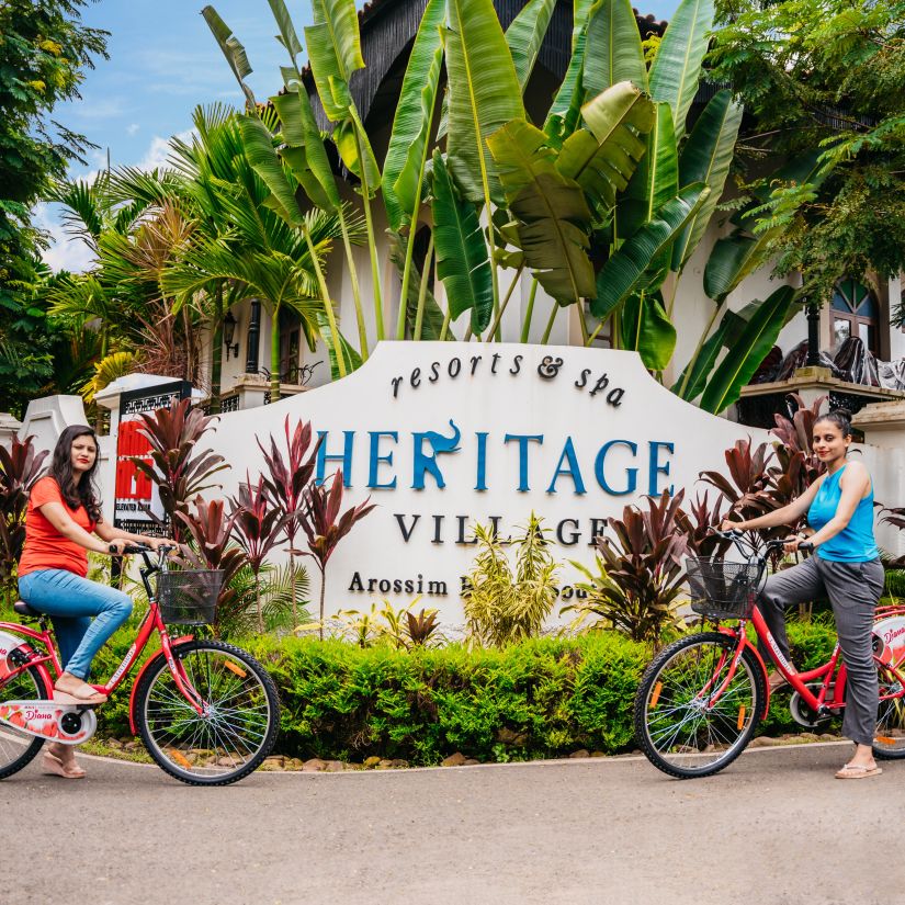 two women on bicycles posing against the backdrop of Heritage Village Resorts & Spa, Goa's sigange