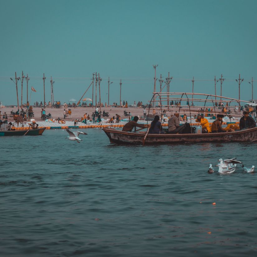  A traditional wooden boat filled with passengers floating on calm waters, with seagulls flying nearby and a crowded beach in the background.