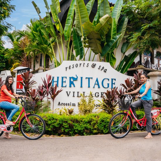 two women on bicycles posing against the backdrop of Heritage Village Resorts & Spa, Goa's sigange