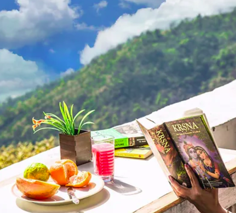 a person reading a book on a balcony with fruits and juice laid out in froint of them