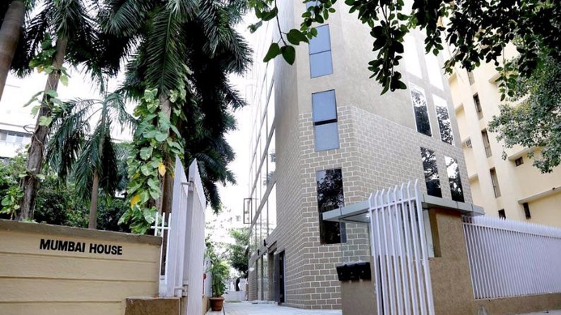 A modern multi-story building labeled MUMBAI HOUSE with a brick facade and reflective windows, flanked by lush green palm trees and a metal gate along a paved pathway.