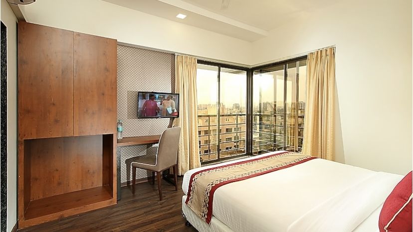 A bedroom featuring a wooden wardrobe, a study desk with a chair, a flat-screen TV, and large windows with curtains, offering an urban view.