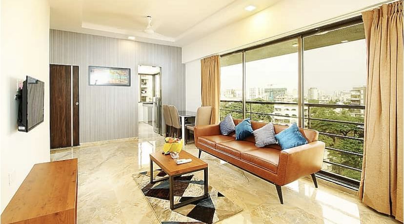 A well-lit living room with a leather sofa, blue cushions, a marble floor, a flat-screen TV on the wall and large windows that offer a view of the city.