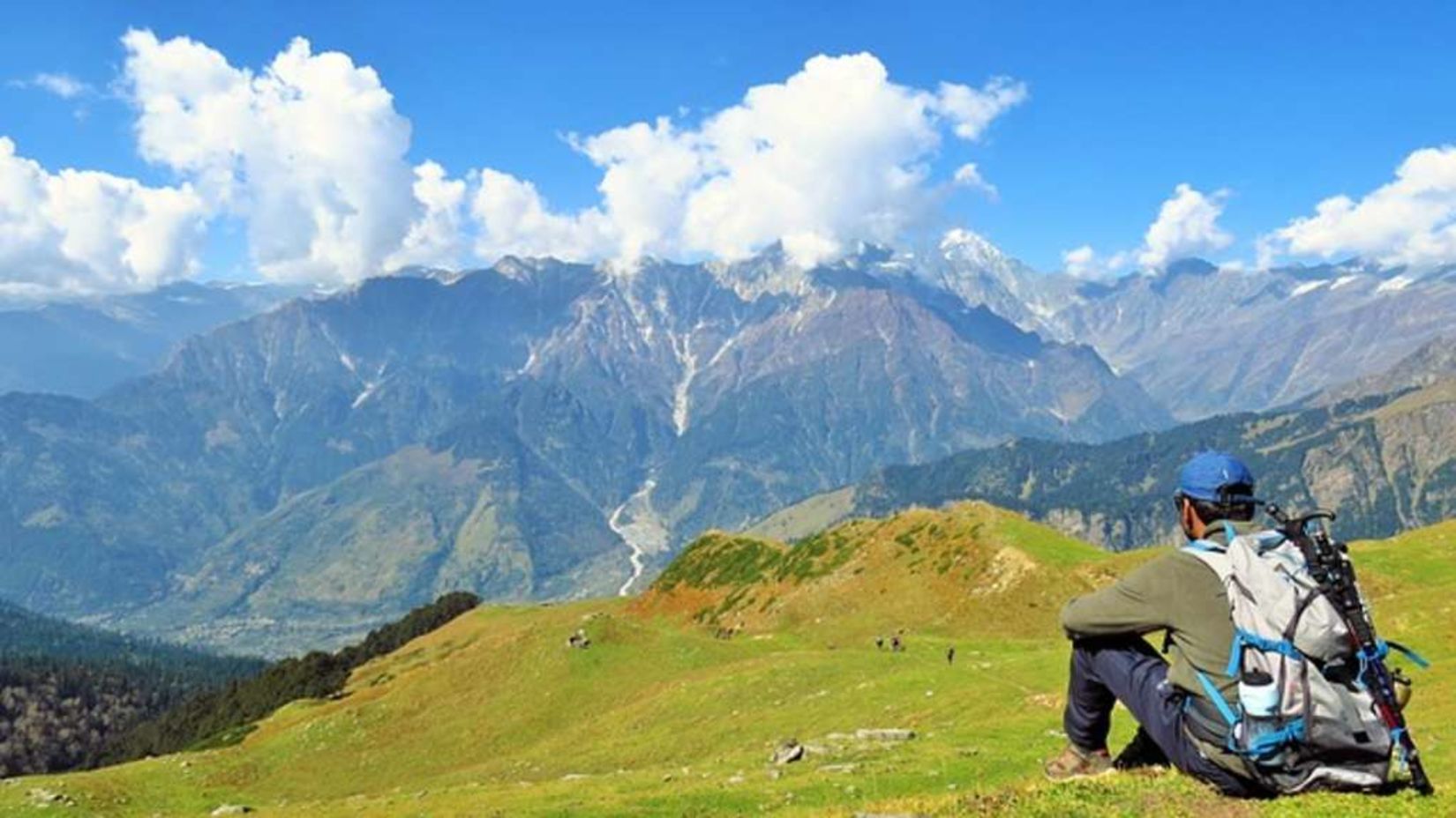 A picture of a man sitting on a hilly terrain with Trekking equipment overlooking the majestic green mountains