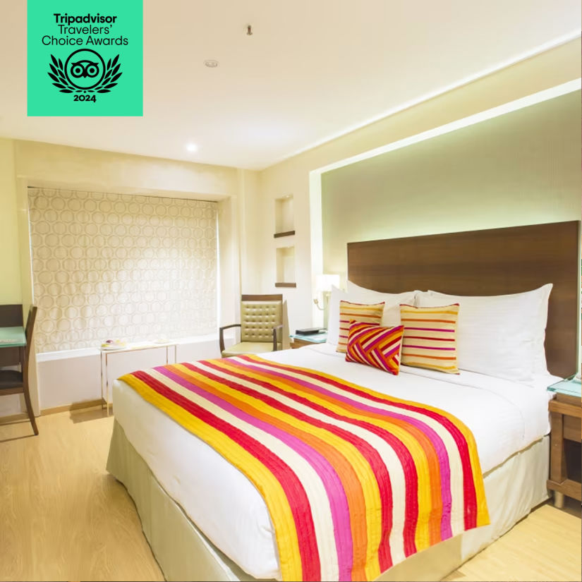 A bright hotel room with a double bed, colorful striped bedding, a window seat, and contemporary decor. Tripadvisor Travelers' Choice Awards 2024 badge displayed.