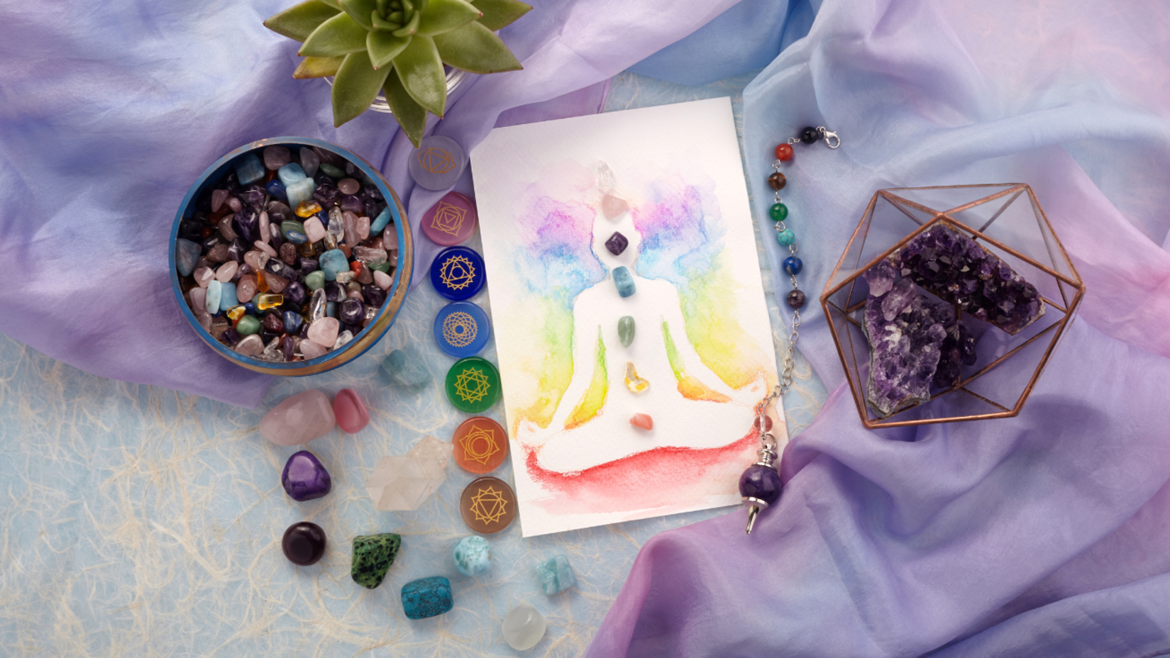 A colorful illustration of a meditating figure with chakras highlighted, surrounded by crystals, gemstones, and a succulent plant on a pastel fabric background.