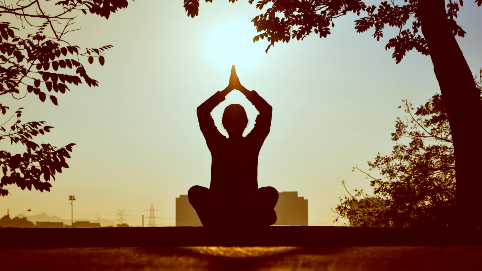 A silhouette of a person sitting cross-legged with hands in a prayer position above their head, meditating outdoors at sunrise or sunset.