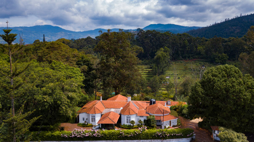 An aerial view of the hotel with trees surrounding it and a tea plantation in the background - Wallwood Garden - 19th Century, Coonoor