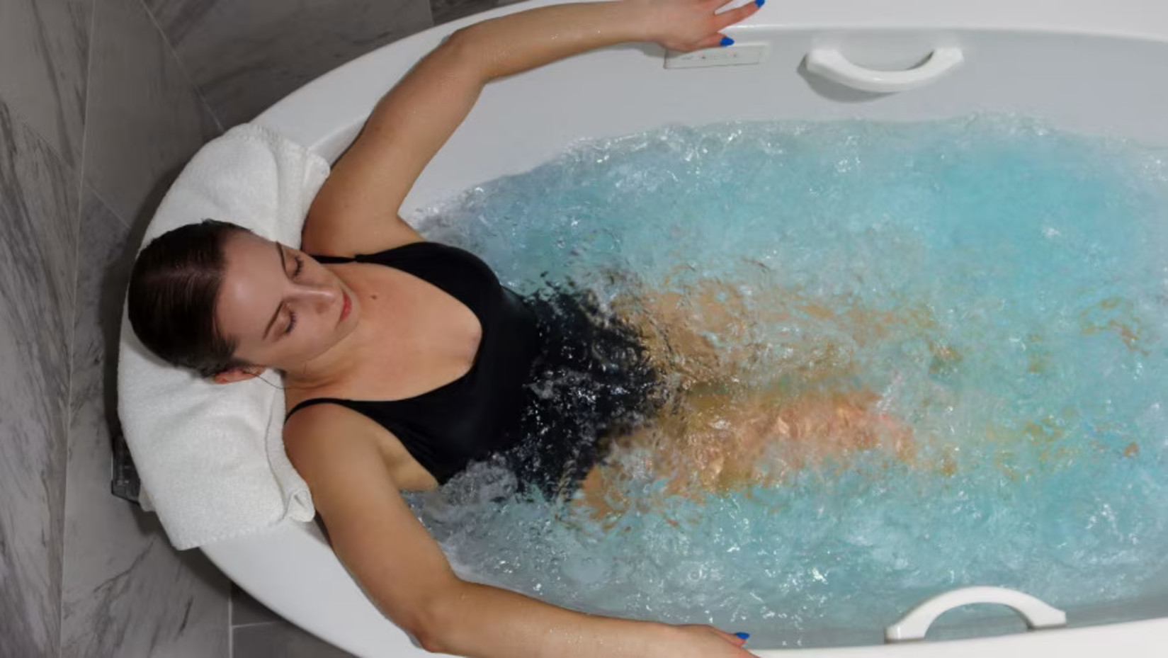 Woman in a black swimsuit enjoying a bubbling hydrotherapy tub, leaning back with eyes closed, against a marble tile background.