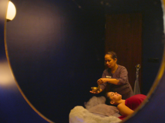 A therapist holding a small bowl and performing a ritual over a client lying on a massage table, reflected in a round mirror.