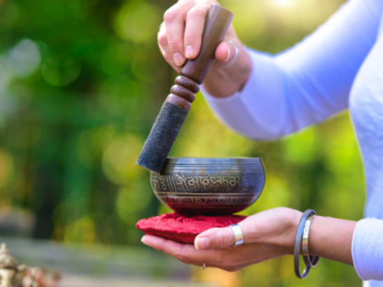Hands holding a Tibetan singing bowl with a mallet, outside against a green, leafy background.