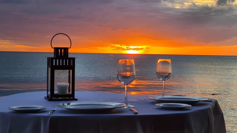 a dinner table set near a beach for a candle light dinner with two wine glasses and captured during sunset