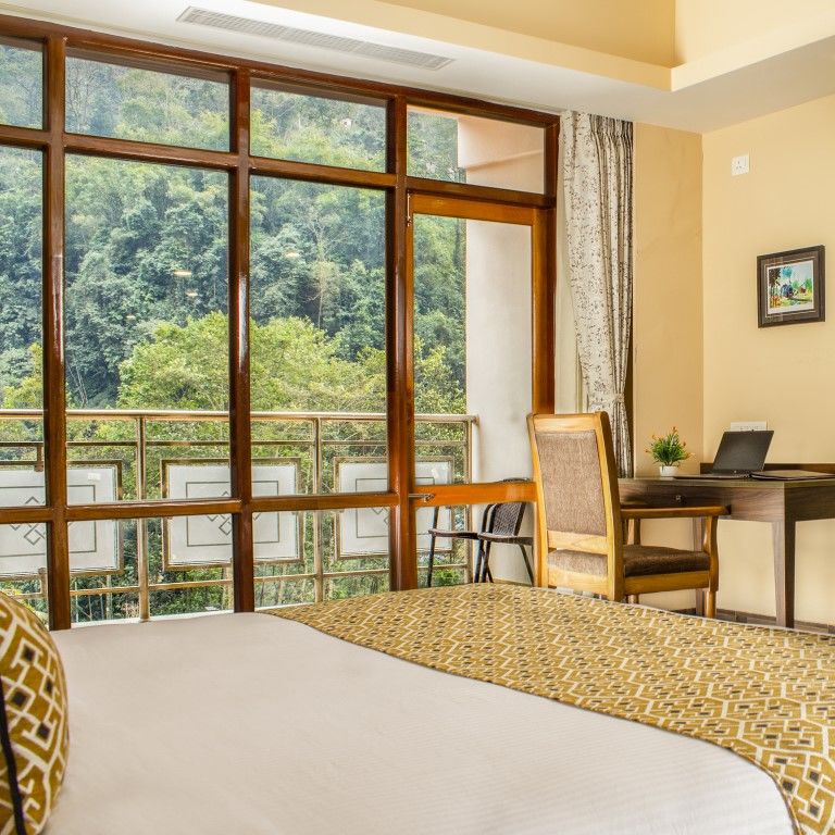 A side view of a bed in a room with glass window, writing desk and a TV on the wall | Sumi Yash Shree Hotels & Resorts