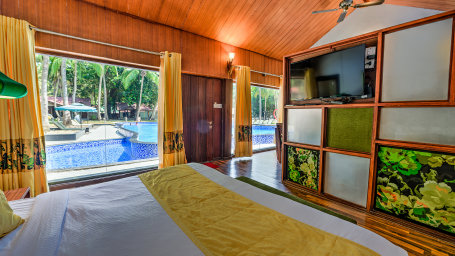 Interior view of a resort bedroom featuring a king-size bed, wooden furniture, tropical artwork, and large windows looking out to a blue pool and palm trees.