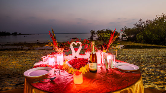 An elegantly set table for a romantic dinner on a beach at dusk, with candles, flowers, and a view of the calm sea.